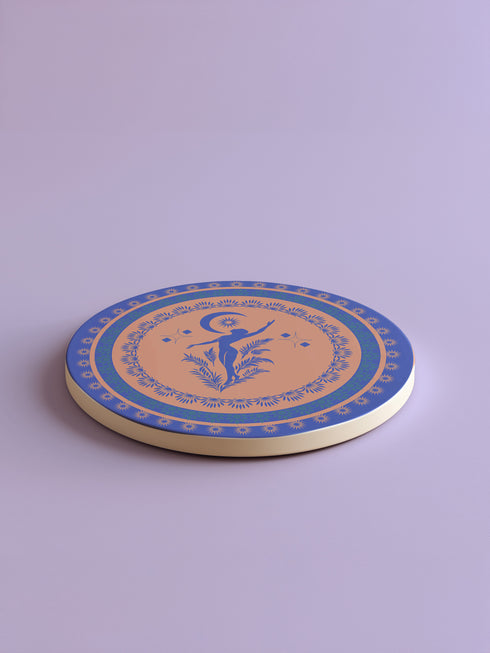 Round ceramic coaster featuring a silhouetted dancing figure reaching toward a crescent moon, surrounded by concentric rings of stars and floral patterns in blue on peach background